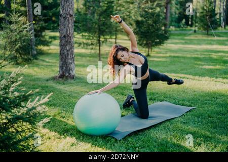Flexible aktive Frau in Sportklothes macht Fitness-Übungen auf Karemat mit Fitball, hebt die Arme und atmet frische Luft im Wald hat Morgenarbeit Stockfoto