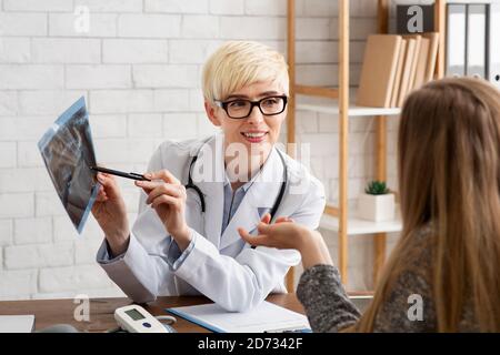 Lächelnde medizinische Frau mittleren Alters in Brille und weißem Mantel Zeigt auf Röntgen und kommuniziert mit dem Patienten Stockfoto