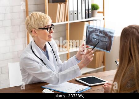 Erwachsene Ärztin in weißem Mantel und Brille zeigt Röntgenaufnahme des Patienten Kiefer und Zähne und Diagnosen, sitzen am Tisch Stockfoto