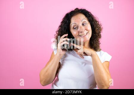 Frau mittleren Alters trägt lässige weiße Hemd stehen über isolierten rosa Hintergrund lächelnd, zeigt auf das Telefon, das er in seinem Ohr hat Stockfoto