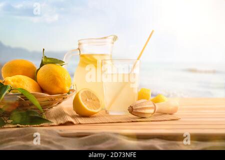 Frühstück mit Krug und Glas gefüllt mit frisch gepresstem Zitronensaft mit Obst auf Holzsockel am Strand. Vorderansicht. Horizontale Zusammensetzung. Stockfoto