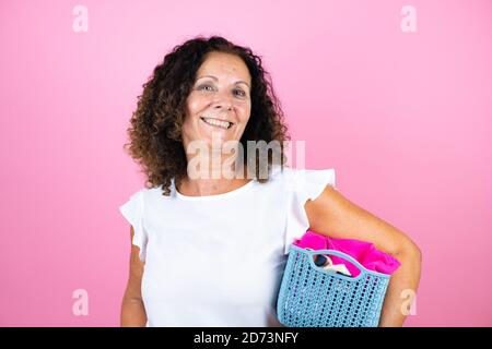 Frau mittleren Alters trägt lässige weiße Hemd stehend über isoliert Rosa Hintergrund tun Hausarbeit halten Korbkorb mit Kleidung Stockfoto