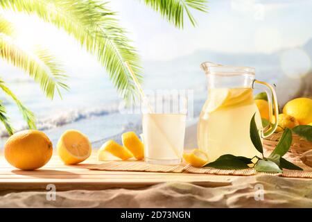 Krug und Glas gefüllt mit frisch gepresstem Zitronensaft mit Obst auf Holzsockel am Strand. Vorderansicht. Horizontale Zusammensetzung. Stockfoto