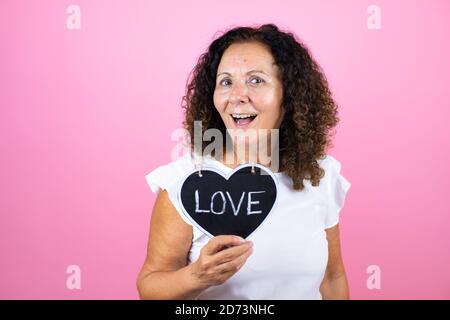 Frau mittleren Alters trägt lässige weiße Hemd stehend über isoliert Rosa Hintergrund lächelnd und halten Herz Tafel mit Liebe Wort Nachricht Stockfoto