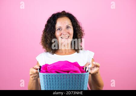 Frau mittleren Alters trägt lässige weiße Hemd stehend über isoliert Rosa Hintergrund tun Hausarbeit halten Korbkorb mit Kleidung Stockfoto