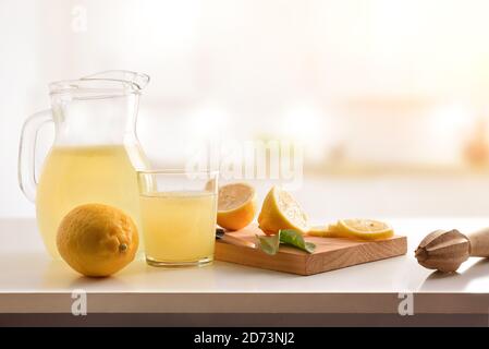 Kanne und Glas gefüllt mit frisch gepresstem Zitronensaft mit geschnittenem Obst auf Schneidebrett auf Küchenbank. Vorderansicht. Horizontale Zusammensetzung. Stockfoto