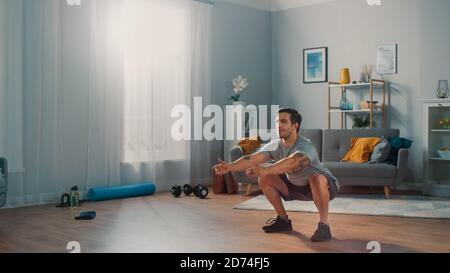 Muscular Athletic Fit man in T-Shirt und Shorts macht Squat Übungen zu Hause in seinem geräumigen und hellen Apartment mit minimalistischem Interieur. Stockfoto