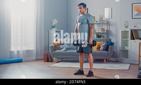 Muskulös Athletic Fit Mann in T-Shirt und Shorts tut Übungen mit Hanteln zu Hause in seinem geräumigen und hellen Wohnzimmer mit minimalistischen Stockfoto