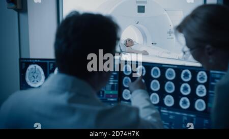 Im Kontrollraum besprechen Arzt und Radiologe die Diagnose während der Überwachung des Verfahrens und Monitore, die Ergebnisse von Hirnscans im Hintergrund des Patienten zeigen Stockfoto