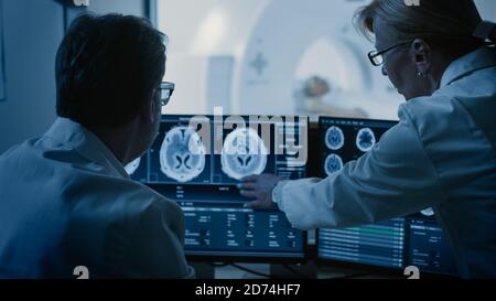 Im Kontrollraum besprechen Arzt und Radiologe die Diagnose während der Überwachung des Verfahrens und Monitore, die Ergebnisse von Hirnscans im Hintergrund des Patienten zeigen Stockfoto