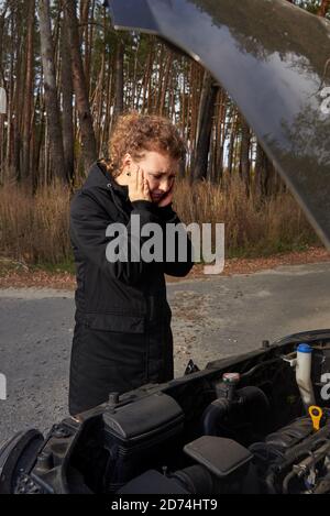 Eine verärgerte Frau steht im Herbst in der Nähe ihres kaputten Autos auf der Straße. Stockfoto