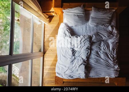 Bett mit weißem Bettzeug in der Nähe des Fensters im rustikalen Holzhaus, Blick von oben. Stockfoto