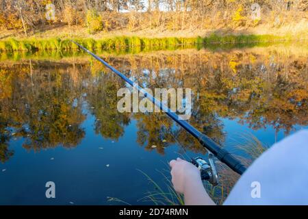 Hand der Fischerin mit Angelrute und Spinnrolle am Flussufer. Konzept der Fischerei als Sport und Hobby. Selektiver Fokus, Herbstlandschaft. Stockfoto