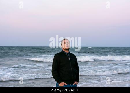 Am Abend steht der junge Kaukasusmann am Strand. Große Wellen mit weißem Schaum bei Sonnenuntergang. Selektiver Fokus. Stockfoto