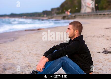 Junger Kaukasischer mit Bart sitzt auf Sand der Küste bei Sonnenuntergang. Selektiver Fokus. Stockfoto