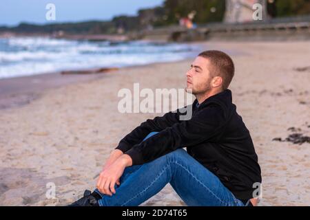 Der junge Mann sitzt abends im Profil an der Küste und schaut auf das Wasser. Romantische Stimmung, selektiver Fokus. Stockfoto