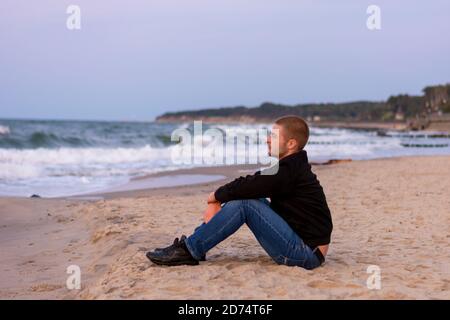 Der junge Mann sitzt an der Küste und schaut mit weißem Schaum auf die großen Wellen. Romantische Stimmung, selektiver Fokus. Stockfoto