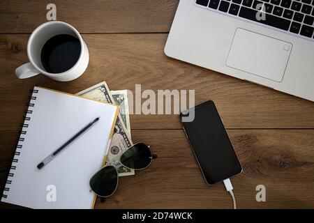 Blick von oben auf den Holzschreibtisch mit Objekten. Laptop, Handy, Notizblock, Kaffeetasse, Geld, Stift und Sonnenbrille. Stockfoto