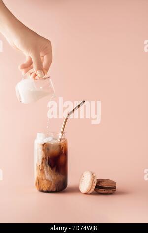 Junge weibliche Hand Gießen Milch in Glas von Eiskaffee in hohen Glas mit goldenem Stroh. Kaffee und Makronen Schokolade, Vanille auf pastellfarbenem Hintergrund für Ihre Gestaltung. Essenskonzept im Vintage-Stil. Speicherplatz kopieren. Nahaufnahme. Stockfoto