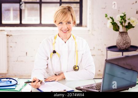 Portrat Aufnahme von lächelnder Ärztin, die am Schreibtisch hinter ihrem Laptop sitzt, während sie im Büro des Arztes arbeitet. Stockfoto