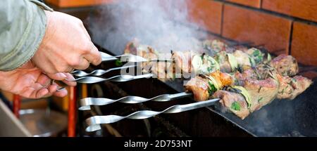 Auf dem Grill wird viel Fleisch auf Spieße geröstet, der Koch dreht seine Shish-Kebabs um. Die Hand eines Mannes dreht sich, fries leckeres Schweinefleisch auf Spieße. Geringe Schärfentiefe. Stockfoto