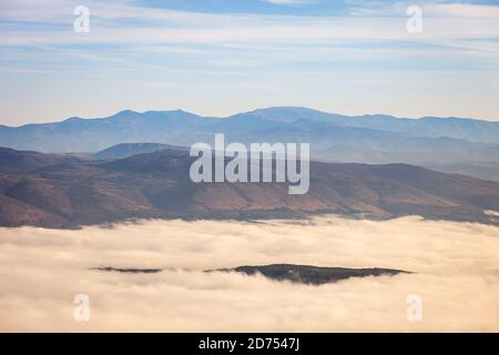 Der Hügel erhebt sich über dem dichten Nebel, der von Wald und fernen, beeindruckenden Bergen am Horizont bedeckt ist Stockfoto