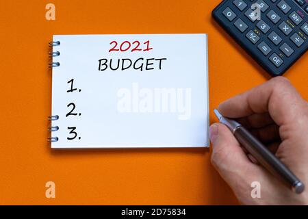 Weiße Note mit Aufschrift '2021 Budget' auf schönem orangefarbenen Hintergrund. Männliche Hand mit Metallstift und Taschenrechner. Geschäftskonzept, Kopierbereich. Stockfoto