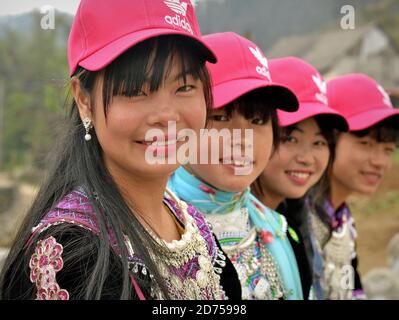 Vier vietnamesische Hmong Hill-Tribe Teenager-Mädchen sitzen in einer Reihe, tragen moderne rosa Baseballmützen und traditionelle Tribal-Kleidung, posieren zusammen für die Kamera. Stockfoto