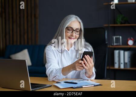 Ältere grauhaarige schöne Frau mit Smartphone und lächeln. Glückliche Geschäftsfrau mit Handy-Apps, SMS-Nachrichten, Surfen im Internet Stockfoto