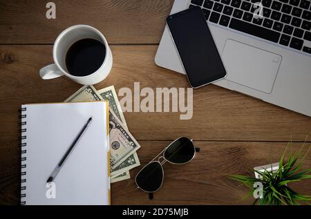 Blick von oben auf den Holzschreibtisch mit Objekten. Laptop, Notebook, Kaffeetasse, Geld, eine Pflanze, Stift und Sonnenbrille. Stockfoto