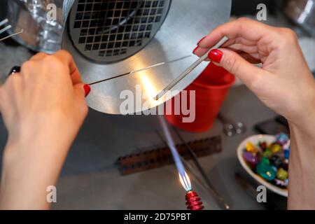 Der Künstler erhitzt das Glas mit einem Gasbrenner. Der Prozess der Herstellung von Glasschmuck. Die Hände des Meisters aus der Nähe Stockfoto