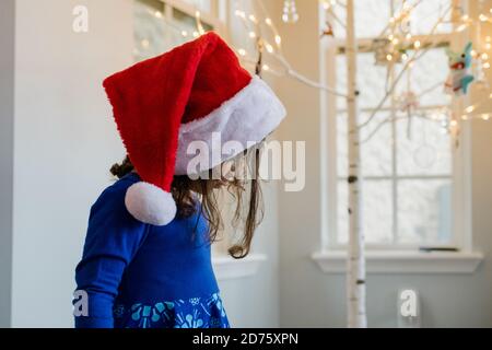 Kleines Mädchen mit Weihnachtsmann-Hut, das vor dem Weihnachtsbaum steht Stockfoto