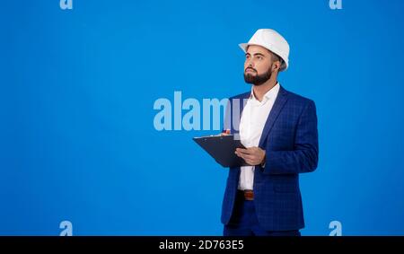 Ein junger Profi trägt einen Hut und trägt einen Zwischenablage auf blauem Hintergrund Stockfoto