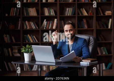 Ein junger bärtiger Geschäftsmann lächelt und schaut in den Rahmen Unter den Büchern in der Bibliothek an einem Tisch in Vorderseite eines Laptops Stockfoto