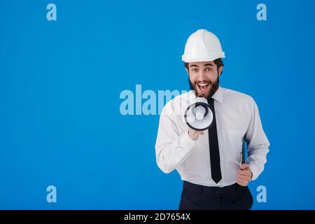 Ein junger bärtiger Bauarbeiter in einem weißen Hemd und Bauhelm auf blauem Hintergrund. Ein Wirtschaftsingenieur-Architekt schreit Stockfoto