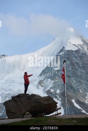 Männlicher Tourist mit Blick auf schöne Berglandschaft und zeigt mit dem Finger. Mann, der den Gipfel erreicht, steht auf Steinen in der Nähe des Fahnenmastes mit Schweizer Flagge. Natur mit herrlicher Aussicht. Sporttourismus in den Alpen. Stockfoto