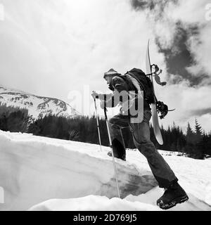 Bergsteiger beim Skilanglauf auf dem schneebedeckten Bergrücken mit Snowboard und Rucksack auf dem Rücken hinauf. Low-Angle-Ansicht. Weiße Wolken am Himmel, Schnee- Stockfoto