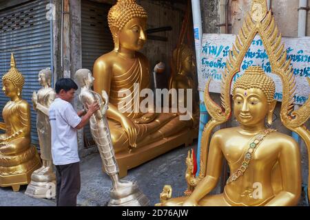 Neben mehreren fertigen Buddha-Statuen, ein Mitarbeiter einer Buddha-Fabrik bewegt eine Statue für einige letzte Arbeit; Bamrung Muang Rd., Bangkok, Thailand Stockfoto