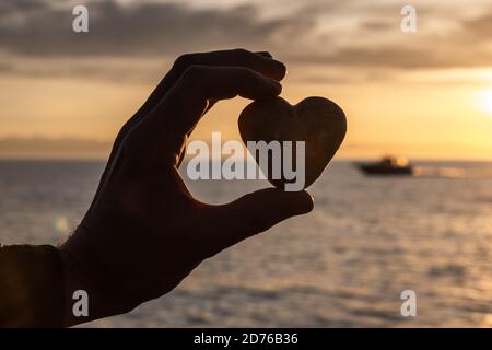 Silhouette eines Mannes Hand hält ein Herz vor dem Hintergrund eines Sonnenuntergangs auf dem Meer, mit einem Schiff segeln im Hintergrund. Valentinstag-Konzept. Stockfoto
