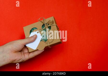 Karton mit Geschenk und Hand auf rotem Hintergrund. Hand des Mannes hält verpackte Geschenkbox. Braune Pappschachtel mit Garn gebunden. Stockfoto