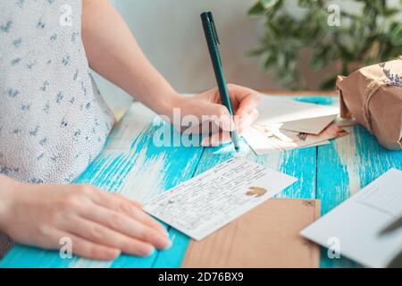 Eine Frau schreibt einen Brief auf eine Postkarte. Hände, die einen Stift in Nahaufnahme halten. Blauer Holztisch mit einem Paket im Hintergrund. Das Konzept der Postcrossing. Stockfoto