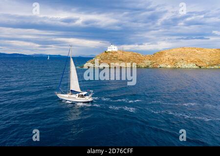 Segeln. Segelboot mit weißen Segeln, welligen Meer und bewölktem Himmel Hintergrund, Leuchtturm auf einem Kap. Griechenland, Insel Kea Tzia. Sommerferien in der Ägäis se Stockfoto