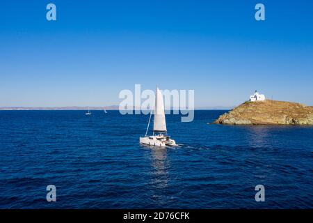 Segeln. Segelboot-Katamaran mit weißen Segeln, geriffelte Meereshintergrund, Leuchtturm auf einem Kap. Griechenland, Insel Kea Tzia. Sommerurlaub in Ägäis. Ae Stockfoto