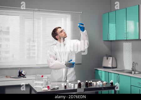 Ein Wissenschaftler in Schutzbrillen und weißen Overalls, der am Tisch im Wissenschaftslabor steht und neue Substanzen untersucht. Stockfoto