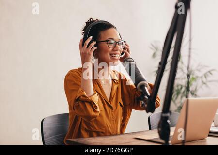 Frau mit Kopfhörern bereitet sich auf die Aufnahme eines Podcasts aus dem Heimstudio vor. Frau, die von zu Hause aus mit einem Laptop und einem Mikrofon auf dem Tisch arbeitet. Stockfoto