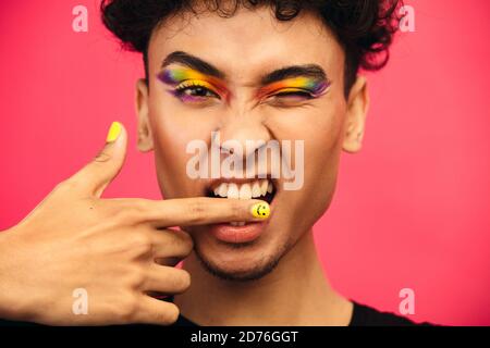 Nahaufnahme eines Transgender-Mannes, der seinen Finger beißt und ein Auge zuzwinkst. Geschlecht Flüssigkeit Mann trägt regenbogenfarbenen Lidschatten und Smiley Gesicht Fingernagel. Stockfoto