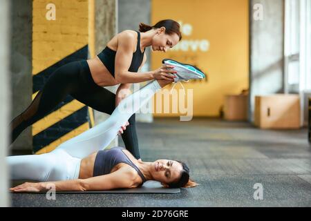 Flexibles Gehäuse. Junge schöne kaukasische Frau, die im Fitnessstudio auf einer Yogamatte liegt und Stretching-Übungen mit Unterstützung des weiblichen Personal Trainers macht. Sport und gesundes Lifestyle-Konzept Stockfoto
