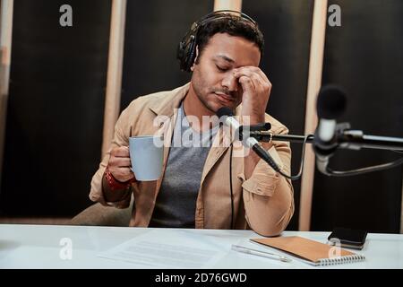 Portrait eines jungen männlichen Radiohosts in Kopfhörern, der müde, erschöpft und Kaffee trinkt, während er sich für die Ausstrahlung im Studio vorbereitet Stockfoto