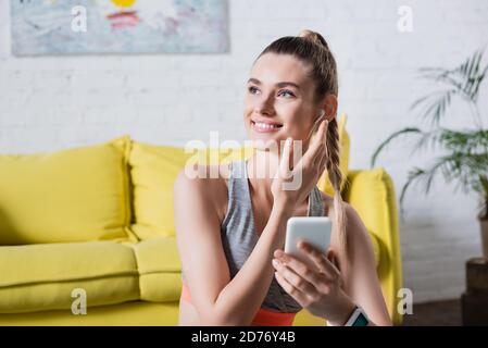 Sportlerin mit Mobiltelefon, das wegschaut und schnurlosen Kopfhörer anfasst Zu Hause Stockfoto