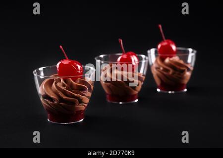 Molkerei Schlagsahne braune Schokoladencreme und rote Marmelade mit frischen Kirschen auf der Oberseite in drei kleinen Gläsern serviert dekoriert. Nahaufnahme von süßen Desserts in Reihe isoliert auf schwarzem Hintergrund. Stockfoto